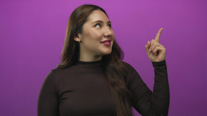Fototapeta premium Woman points her finger upward in purple studio with smiling expression and upward gaze; inspiration.