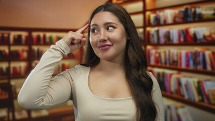 Young hispanic woman pointing head with finger in a well lit library filled with rows of books on wooden shelves; curiosity.