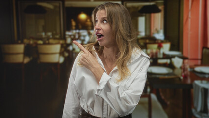 Woman points finger to a table in a restaurant building while leaning forward with open mouth; surprise discovery.