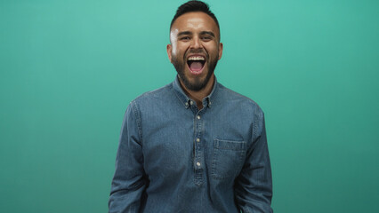 Young hispanic man wearing denim shirt shouting with hand cupped to mouth in teal studio; joy energy.