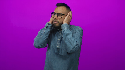 Man covering ears with hands in studio against bright magenta purple wall wearing denim shirt and...