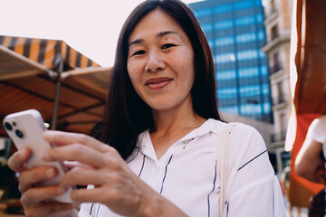 Asian woman smiles gently at camera while holding smartphone, conveying self-confidence, calmness and balance of digital lifestyle in vibrant urban atmosphere.