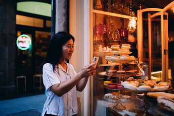 Smiling Asian woman takes smartphone photo of colorful desserts through bakery window, capturing sweet moments and vibrant treats with mobile device while exploring urban life.