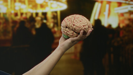 Man holding brain model at outdoor carnival with blurred bright lights, showcasing an educational theme amid a lively fair atmosphere, focusing on human knowledge.