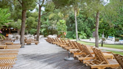 Blurred image of a tropical luxury resort with wooden lounge chairs on a sunlit deck surrounded by lush greenery and palm trees, creating a serene outdoor atmosphere.