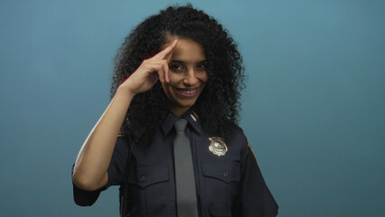 Woman police officer in uniform raising finger salute to head against a blue studio wall; pride...