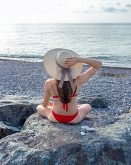 Attractive woman with long hair, wearing red bikini and straw hat, on the beach, sitting on a large rock, sunbathing. Holiday
