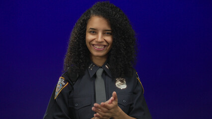 Woman police officer smiling broadly and clapping hands in studio before blue wall with uniform...