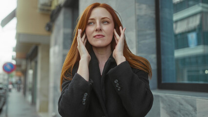 Fototapeta premium Woman in outdoor city setting holds head with closed eyes on street showing discomfort or headache with red hair wearing black coat beside urban building background