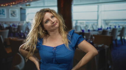 Woman with hands on hips and a sideways glance in a building restaurant, wearing a blue dress and tousled blonde hair; mild annoyance.