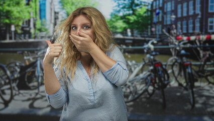 Fototapeta premium Woman with hand covering mouth and thumb pointing back on street by amsterdam canal with bicycles and brick buildings; surprise.