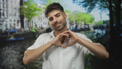 Young hispanic man wearing white shirt makes heart shape with hands on street; affection connection...