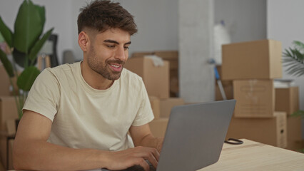 Man typing on laptop in building surrounded by shipping boxes and paperwork spread on table; calm concentration.