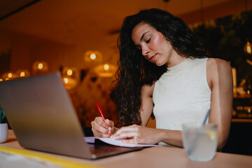 Young female freelancer writes with concentration in front of laptop, embracing creativity and tech synergy in flexible modern working environment.