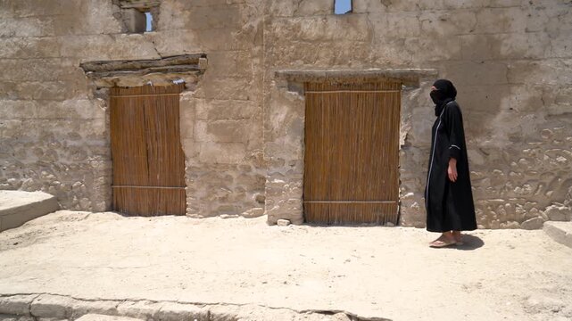 A woman dressed in traditional attire walks gracefully past weathered adobe walls with wooden doors in a historic village on a sunny day, showcasing cultural heritage.
