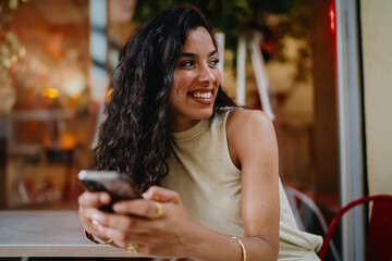 Woman in stylish outfit sits with smartphone at café, symbolizing digital independence and...