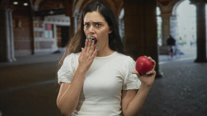 Fototapeta premium Woman holds shiny red apple with extended right hand near brick building; disgust reaction expressed.