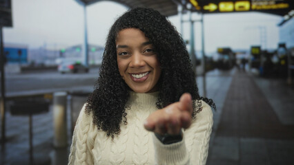 Woman wearing cream sweater extends hand palm up and smiles in busy airport terminal; warm hospitality.