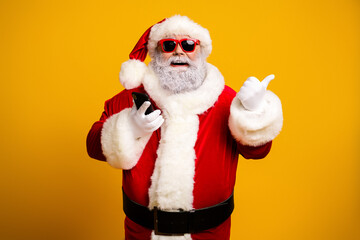 Smiling Santa Claus dressed stylishly in a traditional costume, holding a phone with sunglass on against a yellow background
