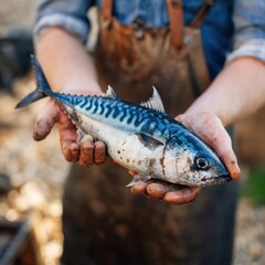 A person wearing an apron and denim shirt holds a freshly caught mackerel with muddy hands.