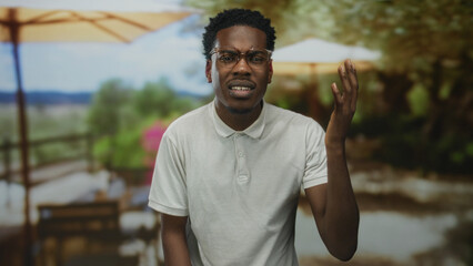 Fototapeta premium Man wearing clear glasses and white polo shirt raises hand palm up at building terrace under sunlight and yellow umbrella in blur; confusion.