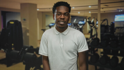 Fototapeta premium Man wearing glasses and a white polo shirt stands smiling by a weight rack in a modern gymnasium; confidence.