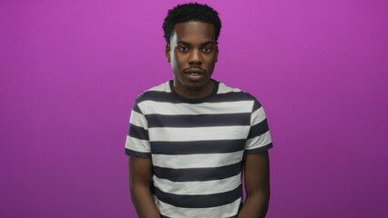 Obraz premium Man in striped shirt winks eye while standing against a vivid purple studio wall with relaxed posture; playfulness.