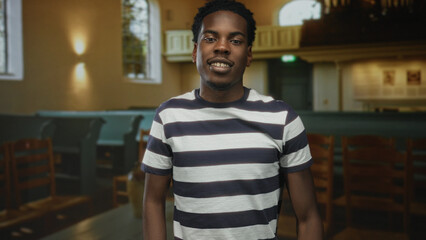 Obraz premium Young african american man in striped shirt gestures thumbs down among wooden pews in church building; disapproval.