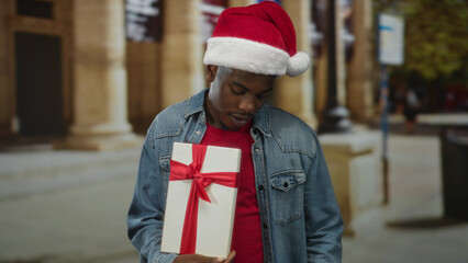 Young african american man wearing santa hat holding gift box and pointing finger confidently on...
