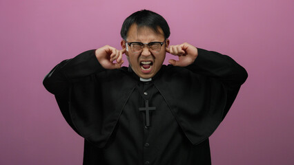 Obraz premium Man in black robe covering ears with hands standing against pink wall expressive gesture emotion glasses religious cross isolated background