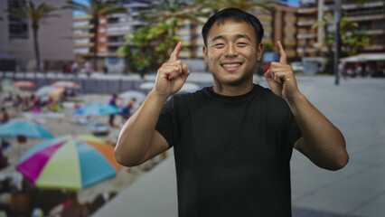 Smiling man pointing both index fingers up wearing a black shirt in front of a beachside building...