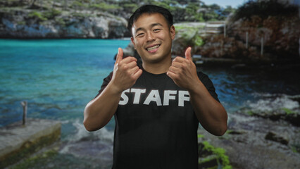 Naklejka premium Cheerful chinese man wearing black staff shirt shows both thumbs up gesture in studio; teamwork and happiness.