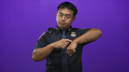 Policeman man pointing finger to bare wrist under his police badge in a purple studio setting; duty.
