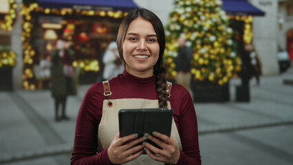 Woman smiling in apron holding tablet in festive decorated outdoor street with blurred christmas tree and storefronts, showcasing holiday spirit and outdoor interaction.