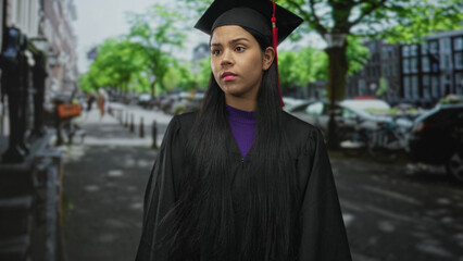 Fototapeta premium Graduate woman wearing mortarboard and gown touches cheek with both hands on street outside building; anxiety.