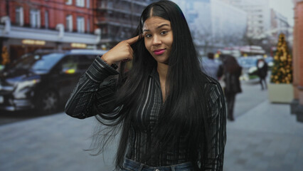 Fototapeta premium Young latin woman in striped shirt and jeans points finger to head in city street amid passing cars and blurred pedestrians; confusion.