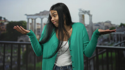 Young latin woman shrugs open palms with puzzled expression in front of ancient building ruins;...