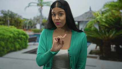 Young latin woman wearing green cardigan pointing finger in building; firm refusal disapproval...