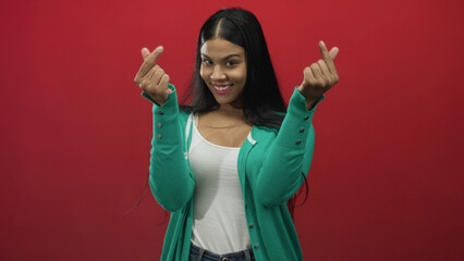 Woman wearing green cardigan and white tank top making finger heart gesture and smiling in studio;...