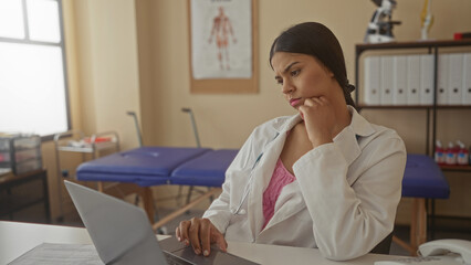 Obraz premium Woman doctor in white coat with hand on chin typing on laptop in clinic building beside anatomy chart; thought concern.