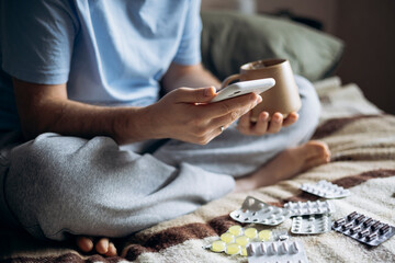 Man with tea and smartphone in bed surrounded by medicine at home.