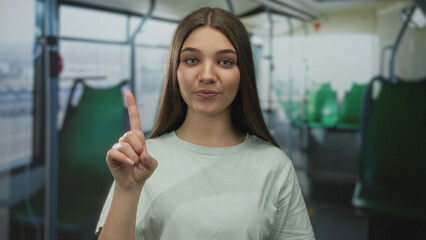 Smiling woman waving open hand inside a city bus with green seats and window view, facing camera; friendly greeting.