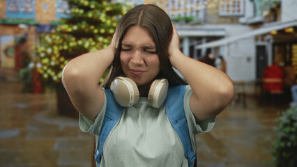 Teen girl with white headphones and blue backpack covers ears on holiday street beside decorated...