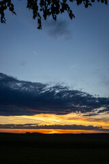 Stimmungsvolle Landschaft bei Sonnenuntergang mit dramatischen Wolken am Himmel, Silhouetten am Horizont und warmen Lichtfarben in Orange und Gelb.