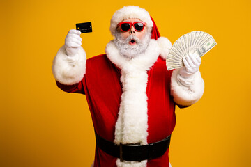 Santa Claus dressed for Christmas holding cash and a credit card on a yellow background, ready for holiday shopping