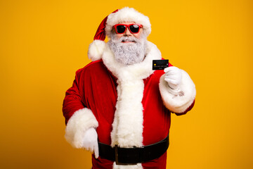 Santa Claus wearing sunglass holding a credit card on a bright yellow background, dressed in his iconic red suit and beard
