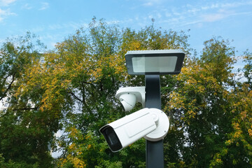 Two white surveillance cameras on a pole with a lantern against a background of trees.