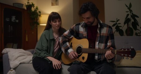 Brunette Guy Uses Tabs To Play Guitar While His Girlfriend Listens To Him In Cozy Apartment At Home In The Evening. Music and guitar evening at home for a couple date - Powered by Adobe
