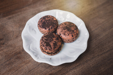 Three bittersweet chocolate ganache macarons with cocoa powder on a white ceramic plate on an old wooden table in cafe dessert shop.