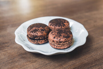 Three bittersweet chocolate ganache macarons with cocoa powder on a white ceramic plate on an old wooden table in cafe dessert shop.